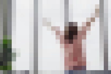 Person stands with arms raised beside a window with vertical bars, suggesting a scene behind bars or a cage-like setting